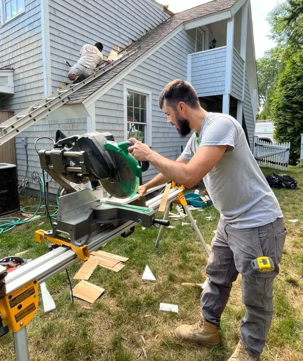 Contractor cutting wood for residential siding repair while another worker installs shingles on a home in Cape Cod